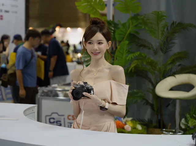 Young woman at a trade show booth holding a camera, surrounded by indoor plants and visitors. Trade show marketing claims: checks for UK stands